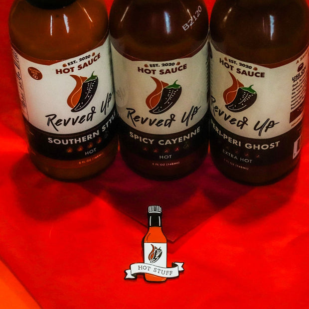 Hot Stuff soft enamel pin with southern style, spicy cayenne, and peri peri ghost hot sauces in the background. background. This hot sauce pin is a spicy way to show off your favorite condiment. Available exclusively here at Revved Up!