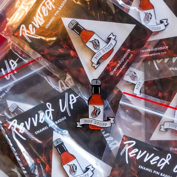 Hot Stuff soft enamel pin with piles of pins as the background. This hot sauce pin is a spicy way to show off your favorite condiment. Available exclusively here at Revved Up!