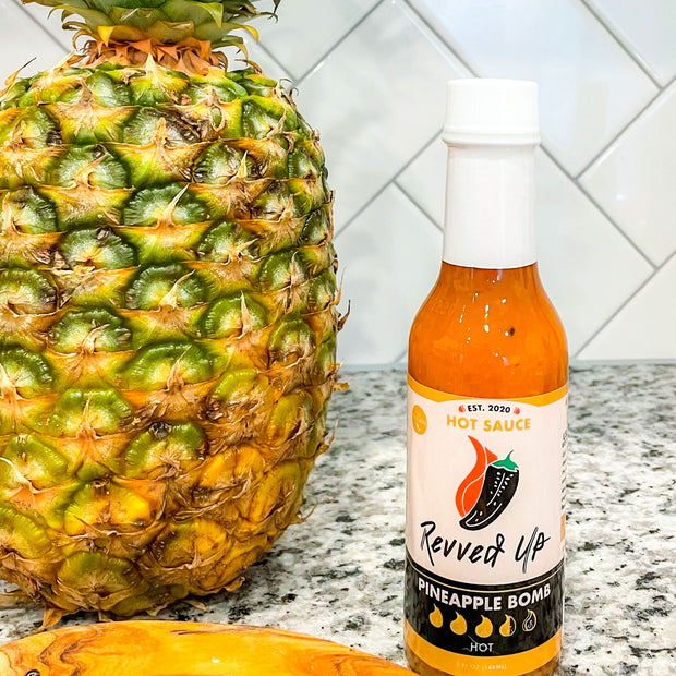 Pineapple Bomb Hot Sauce - | Revved Up. Pineapple Bomb Hot Sauce. Made with real pineapples instead of the artificial stuff typically found in other hot sauces, this zesty mixture gives your boring food new life while giving a tropical sweetness with a medium-high heat that will have your mouth watering for more. Kick up lunches such as chicken sandwiches and burgers or add some flavor to traditional Mexican dishes like burritos and tacos! Pineapple bomb hot sauce in the kitchen next to a pineapple. 